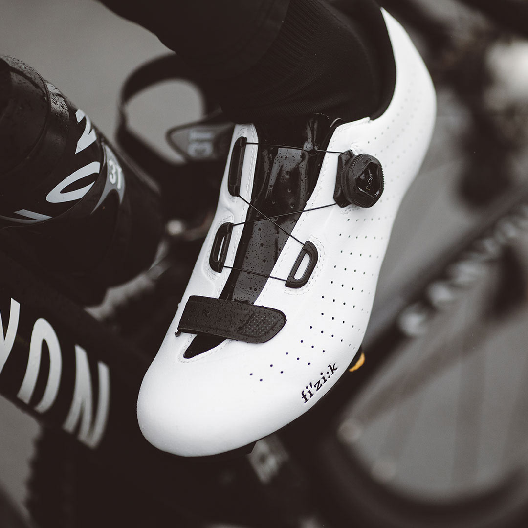 A close up shot of a cyclist's feet. The cyclist is wearing white bicycle shoes by Fizik.