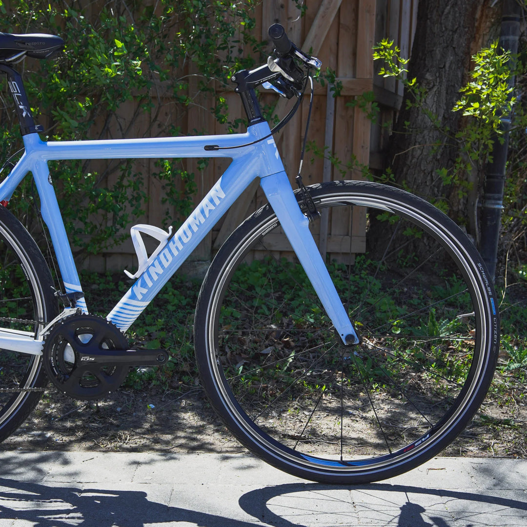 A close-up shot of a light blue hybrid bicycle made by KindHuman Bicycles. The bike is standing on a side walk beside grass and wooden fence.