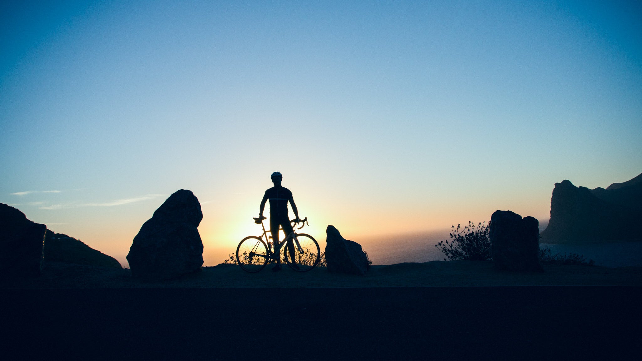 A silhouette of a man standing with a bike in front of a sun rise.